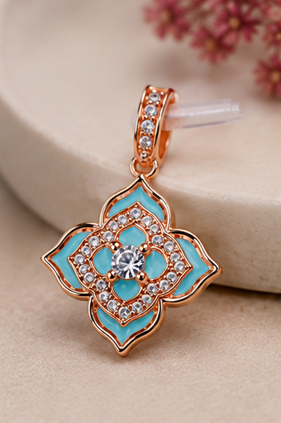 A close-up of a floral pendant charm with turquoise blue enamel inlay and rose gold metal, accented with small clear stones, resting on a beige surface with a soft-focus background.